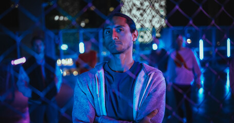 Portrait of a Focused Multiethnic Man Looking at Camera while Standing Behind a Fence in an Urban City Environment at Night with Neon Lights. Stylish Diverse Group of Friends Standing Behind Him