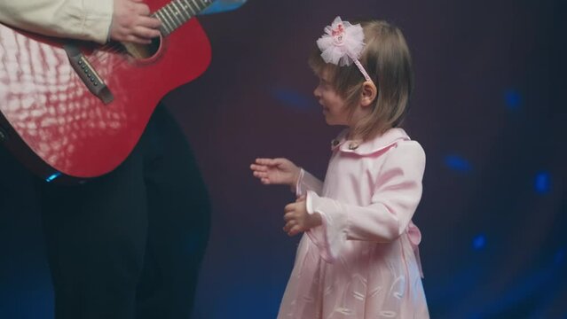 Little girl on stage in pink vintage dress, she dances, her father plays acoustic guitar. Color music is shining and smoke is billowing. Father Day. Performance on stage. Hobbies and creativity