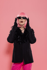 Drag queen in wig and jacket touching sunglasses on pink background.