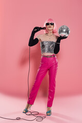 Full length of trendy drag queen in sunglasses and gloves holding microphone and disco ball on pink...