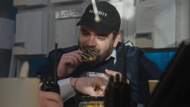 Two security officers in uniform have a lunch break in the room for monitoring surveillance cameras. They eat donuts, talk, discuss work details. Two walkie talkies stand on the table. Slow motion.