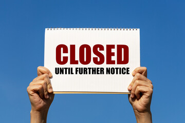 Closed until further notice text on notebook paper held by 2 hands with isolated blue sky background. This message can be used as business concept about closing shop.