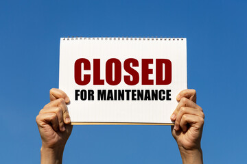 Closed for maintenance text on notebook paper held by 2 hands with isolated blue sky background. This message can be used as business concept about closing shop.