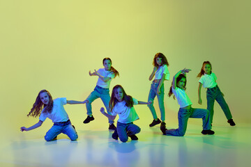 Group of talented, active, little girls, children in casual, sportive clothes dancing hip-hop against green studio background in neon light. Concept of childhood, hobby, sportive lifestyle
