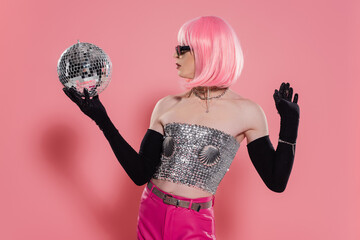Side view of trendy drag queen in sparkling top and gloves holding disco ball on pink background.