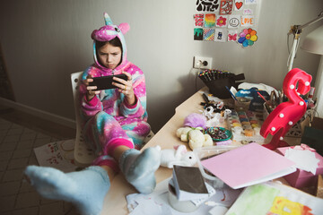 Serious teen girl using phone, playing phone games in messy childrens room, kid playing among the many toys at home, a lot of toys and things scattered on table, dirty home