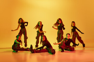 Art. Group of children, little girls in sportive casual style clothes dancing hip-hop, contemp dance against yellow studio background in neon light. Concept of childhood, hobby, sportive lifestyle