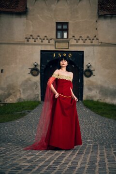 Vertical shot of a Caucasian woman wearing a medieval red dress posing against ancient buildings