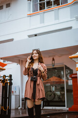 female traveller in brown trench coat calling on phone while standing at front of the bulding