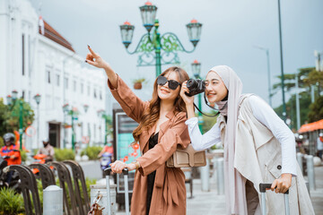 woman with hijab operating her camera while standing beside the woman with long brown hair that pointing with her finger