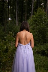 Vertical shot of the pretty woman from behind wearing a long purple dress and standing in the woods