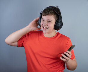 A young man with headphones is happy to listen to music from his phone and smiles happily. A teenager in a red T-shirt and black headphones on a gray-blue background