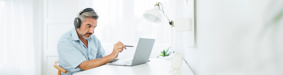 Senior Mature adult Man in headphones having video call on laptop computer, Online Meeting, Working at home, Video conference, Remote Location. Banner, Panoramic.