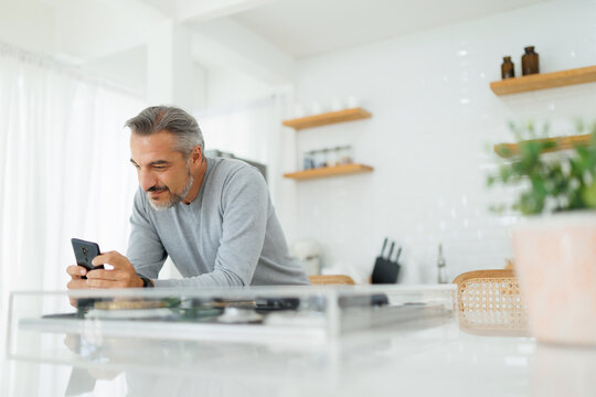 Smiling Mature adult male using smartphone or mobile phone at home. Middle aged Man reading message on the phone in kitchen.