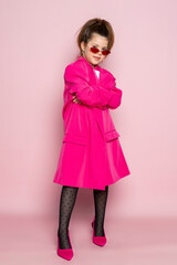 Portrait of a little girl in an oversized pink blazer and a slim stylish red glasses. Child wearing adult clothes. Isolated on pink background.