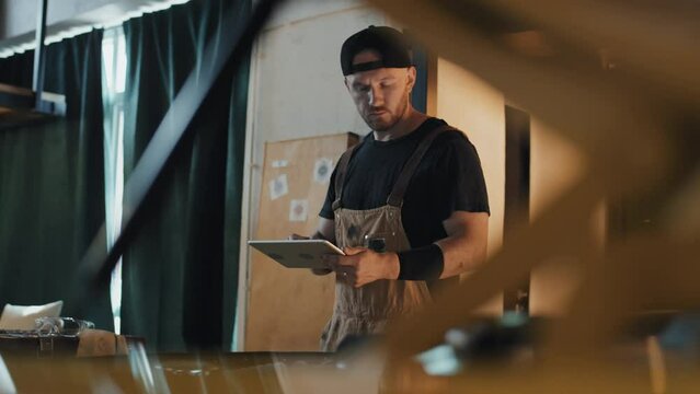 Medium shot of tattoed auto mechanic wearing coveralls and cap making car diagnostics with tablet in his hands, view from under motor hood, parking house
