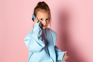 A little girl in an oversized blue blazer. Child wearing adult clothes and talking on the phone. Isolated on pink background.