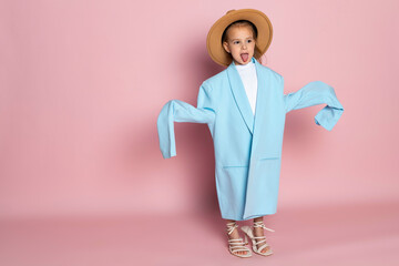 Portrait of a little girl in an oversized adult blue blazer and a round brown hat. Child wearing adults clothes. Isolated on pink background.