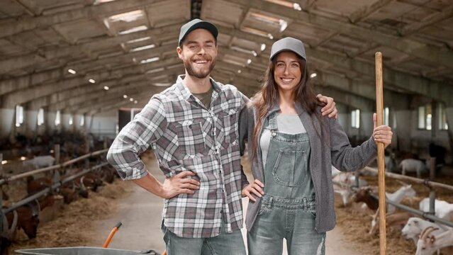 Zooming in pretty Caucasian couple with agricultural tools, standing in goats barn. Happy man hugging his wife and smiling. Young people taking care of farm animals together. Farming business concept.
