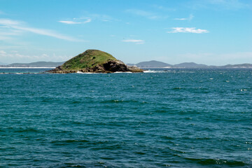 Island seen from Foguetes beach, close to the city of Cabo Frio, with many rocks and waves crashing on them, blue sky and clear sea.