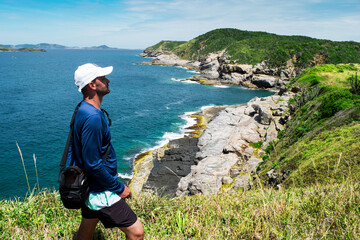 Man enjoying a view from above of the beautiful Praia das Conchas, near the city of Cabo Frio, with blue sea around, undergrowth, large rocks and mountains in the background.