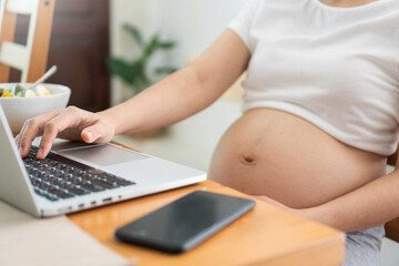 Fototapeta premium Pregnant Woman sitting holding blue credit card and making online payment shopping on laptop computer at home. Female making purchases in the Internet banking. Spending money and lifestyle.