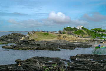 View of Praia do Forte, with the fort on top of the hill, sea, large rocks, cloudy sky, fishing boats and the city in the background.