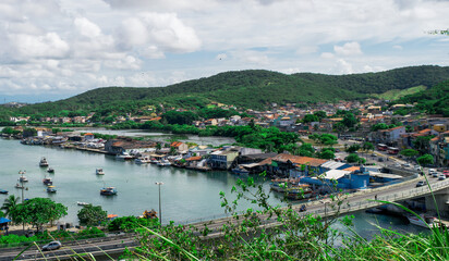 Fototapeta na wymiar View of the port of Cabo Frio seen from the viewpoint of Nossa Senhora da Guia, with a bridge and many boats sailing through the channel, mountains around and a beautiful blue sky with clouds.