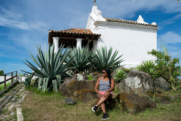 Woman sitting near the chapel at the Nossa Senhora da Guia viewpoint, located in Cabo Frio, stone path, surrounding vegetation and beautiful blue sky.