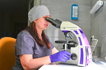 Obraz premium A laboratory female worker in blue gloves looks into a microscope and examines fragments of tissue. Examination under microscope. Test sample under the microscope in laboratory. Microbiology research