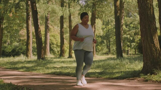 Full length slowmo of body positive mature Black woman jogging on park trail at summertime