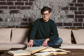 A hard learning student dressed neatly, in round glasses is reading and studying among piles and...