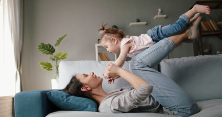 Active caucasian young mom and her little cute daughter playing at home, playing plane on couch. Joyful babysitter working at home - happy family, activity together concept 