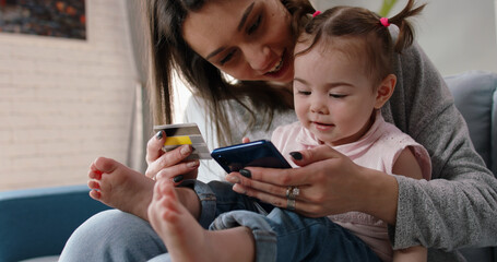 Relaxed caucasian mom is making an online purchase with credit card using smartphone while holding her cute little baby girl on knees - happy family, technology, finance 