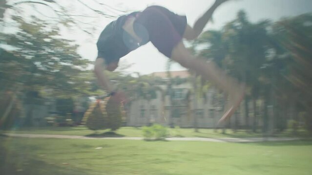 Panning slow mo shot of sporty man running and performing sideflip while practicing parkour stunt in park on summer day