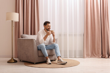 Smiling man drinking tea while resting on armchair near window with beautiful curtains at home....