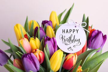 Beautiful bouquet of tulip flowers with Happy Birthday card on light background, closeup