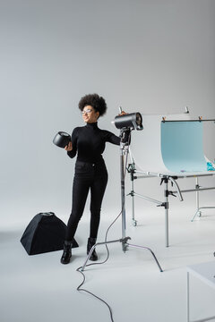 full length of cheerful african american content producer assembling lighting equipment near shooting table in photo studio.