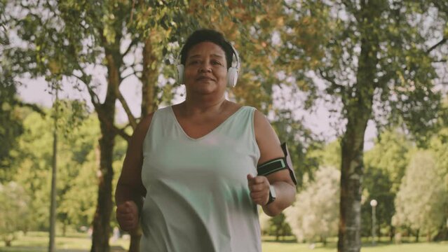 Medium slowmo of happy body positive mature Black woman in wireless headphones sports walking outdoors in park on warm sunny day