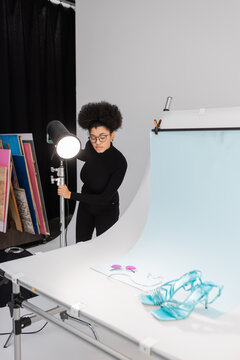 african american content producer in eyeglasses near spotlight and stylish sunglasses with sandals on shooting table in photo studio.