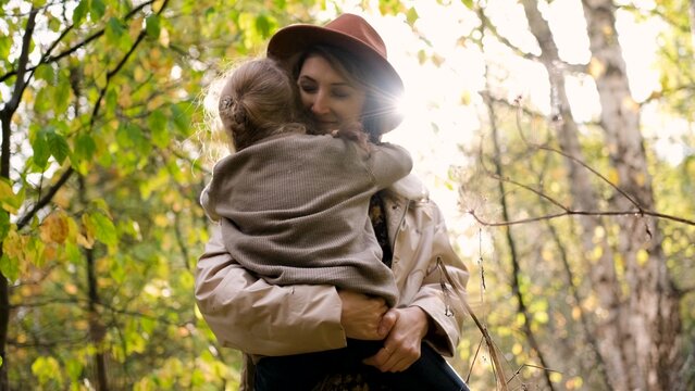 Hello September. A young mother in a brown hat and a little daughter walk in the city park in autumn. Mom holds the girl in her arms and hugs her tightly with love. - Powered by Adobe
