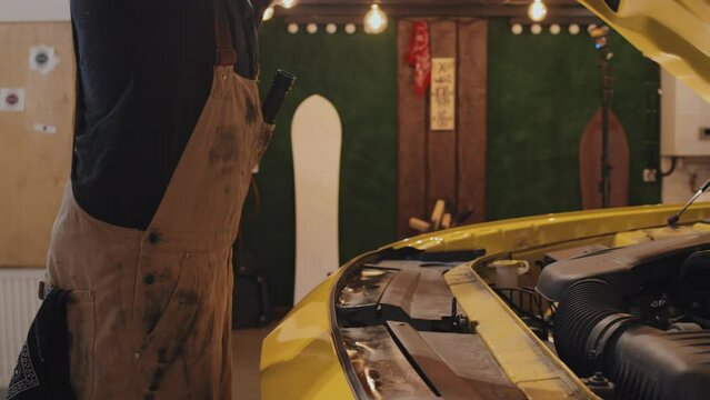 Midsection lateral shot of mechanic wearing coveralls opening yellow motor hood, taking out torch, highlightning interior. Garage wall and light bulbs in background