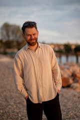 Stylish hipster bearded man with curly hair standing at the lake beach and smiling at the sunset in beige linen shirt