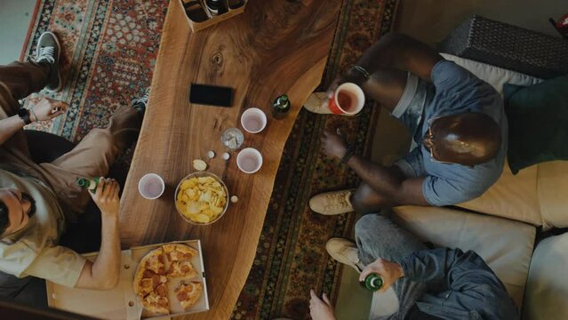 Top shot of three friends, two Caucasian man and one African American man, clinging cup and bottles sitting behind long table with pizza, snacks and phone on it. Floor covered with rug, small party