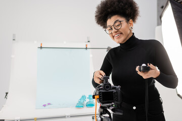 cheerful african american content maker with exposure meter smiling near digital camera in modern studio.