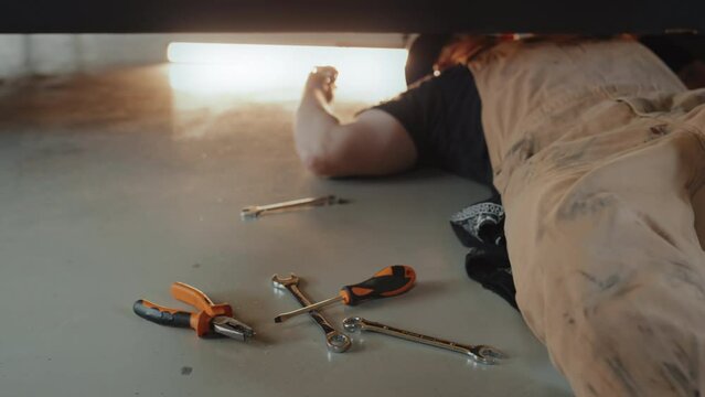 Cropped full medium shot of auto mechanic lying on garage floor, picking tool after tool, wrench and screwdrivers beside him. Mechanic making repairments under car body