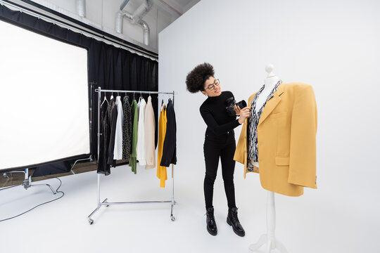 full length of african american content producer with digital camera adjusting trendy blazer on mannequin in photo studio.