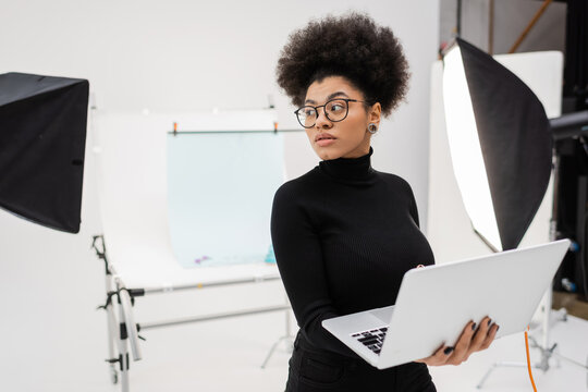african american content manager with laptop looking away near spotlights and blurred shooting table in photo studio.