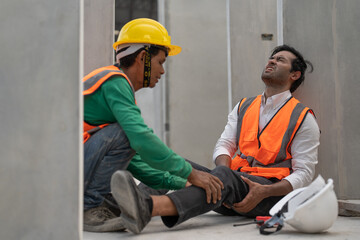 Asian man engineer feeling pain in his leg from accident while working in factory making precast concrete wall for real estate housing. Male worker colleague is helping him.