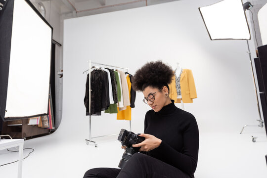 african american content maker looking at digital camera while sitting near trendy clothes in photo studio.